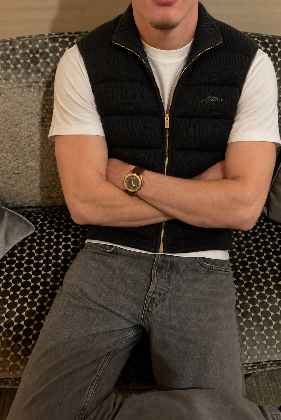Black knitted vest styled with a white T-shirt and grey jeans, seated on a patterned sofa